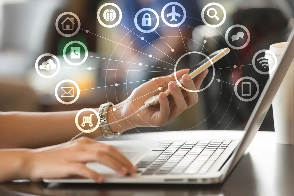 Mobiltelefont tartó kéz egy laptop mellett, körülötte internet-, levelezés- és biztonsági ikonok